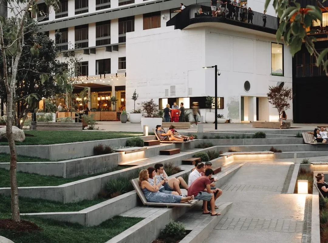A vibrant community space in Hamilton featuring people relaxing on tiered seating, lush greenery, and modern architecture.