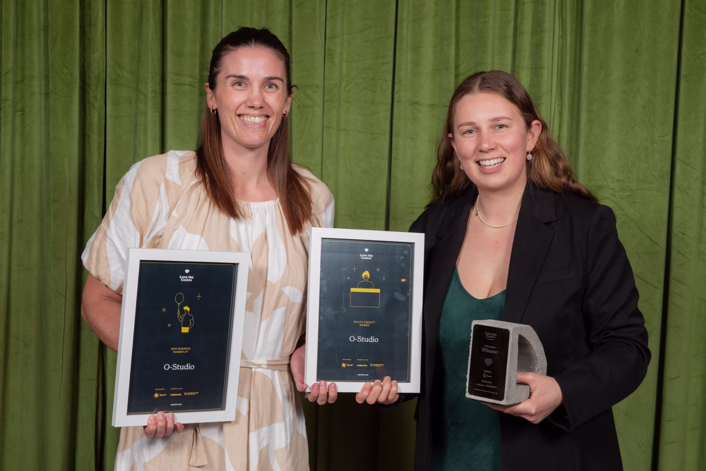 Two women smiling and holding framed awards and a trophy against a green curtain backdrop.