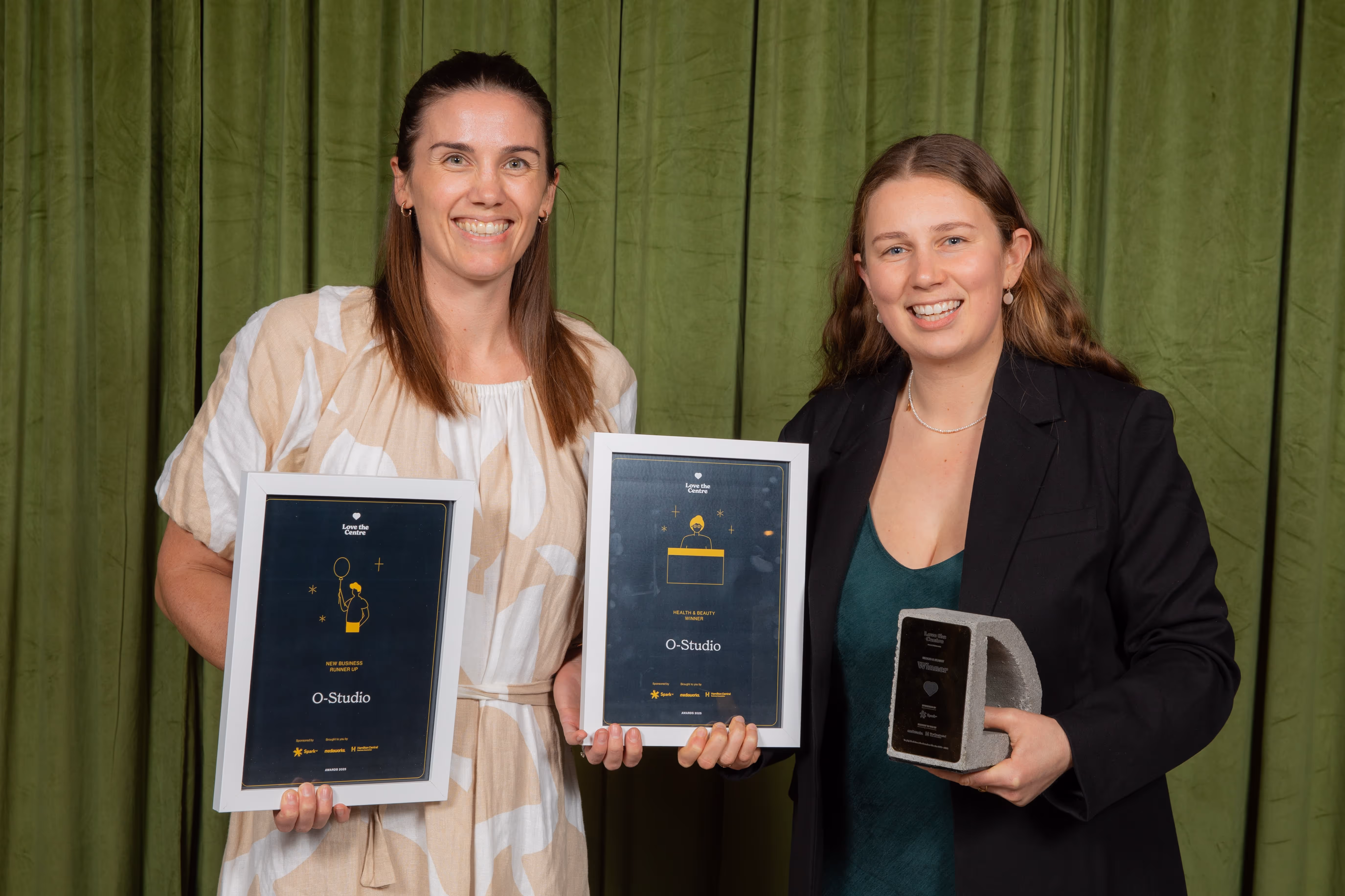 Two women smiling and holding framed awards and a trophy against a green curtain backdrop.
